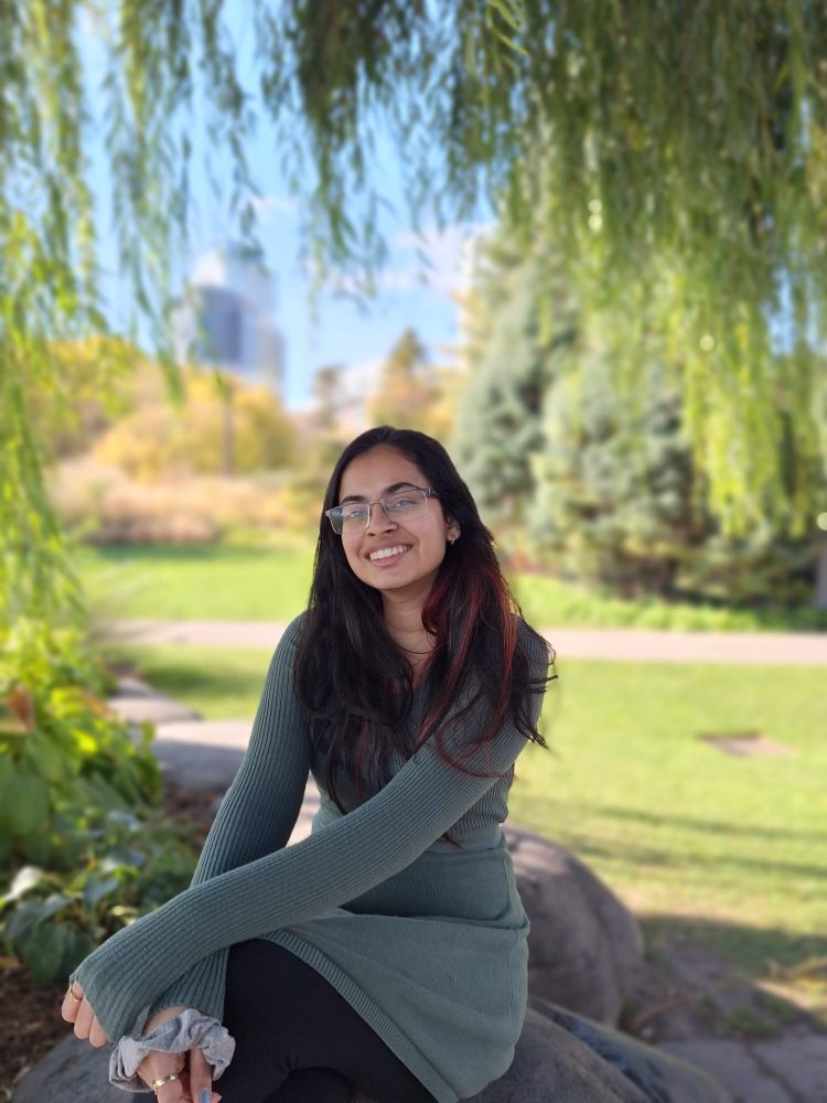 A smiling young woman with long brown hair and glasses sits outside.